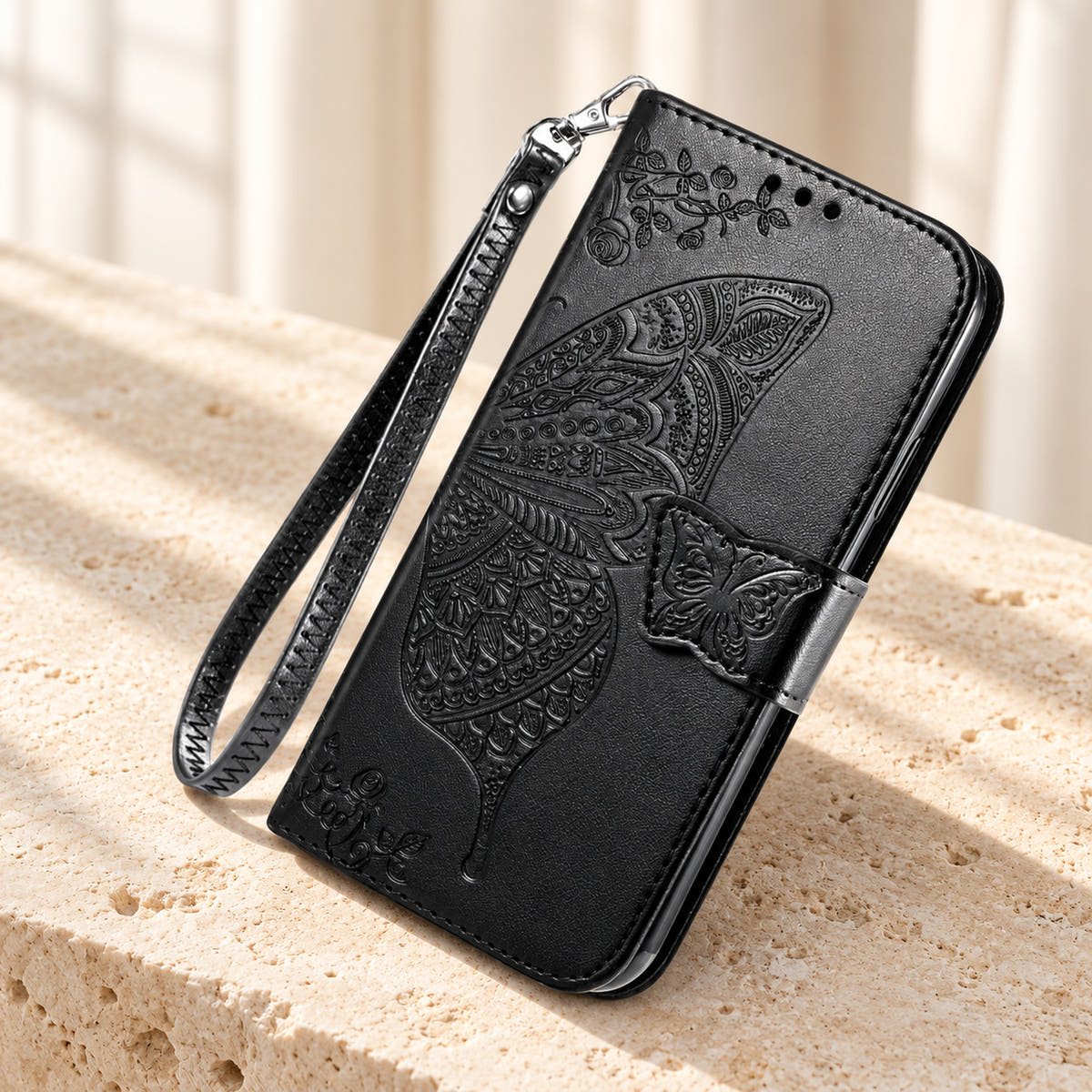 Butterfly embossed iPhone wallet case in Black, tabletop front view showing the detailed wing, tab closure and clip-on wrist loop.