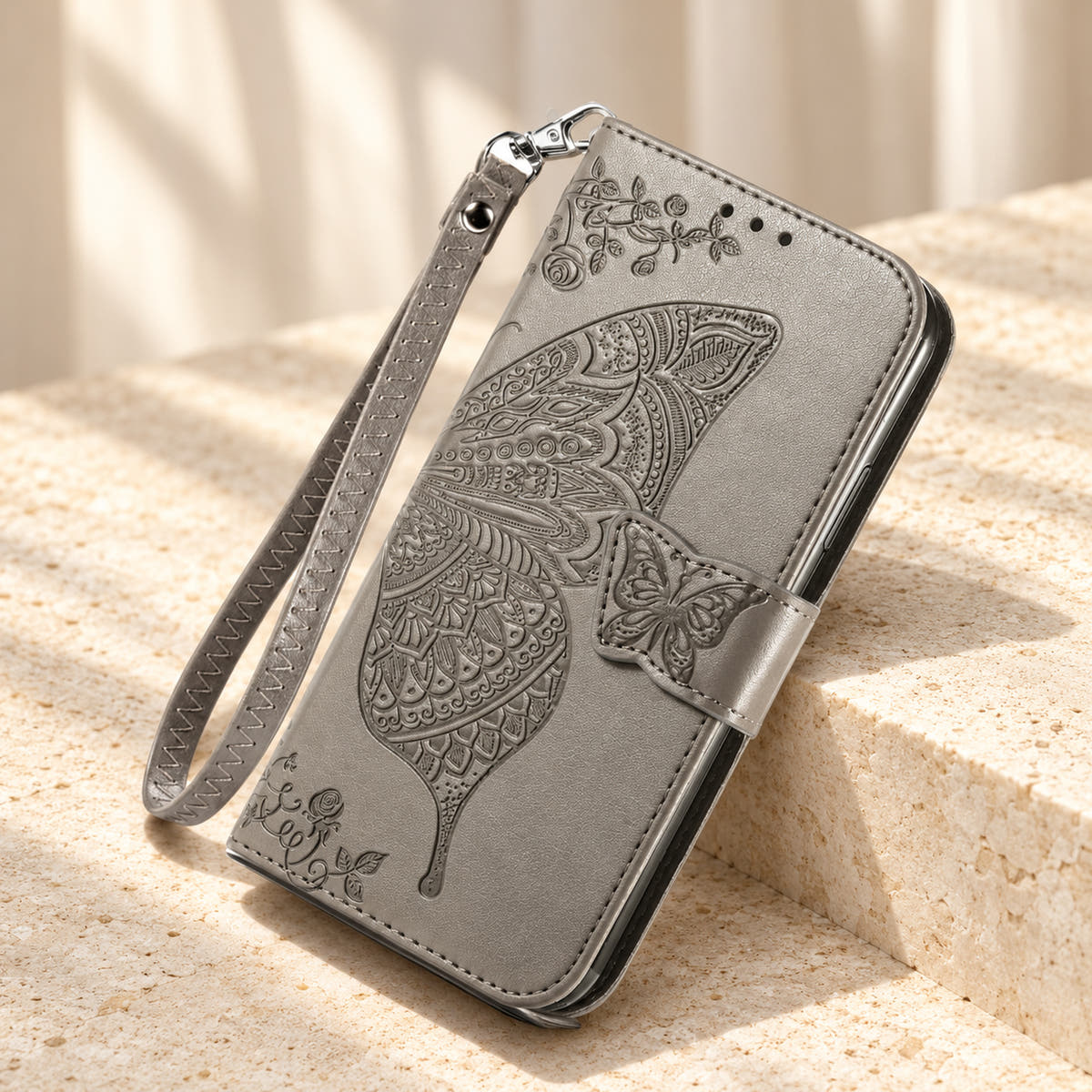 Butterfly embossed iPhone wallet case in Gray, tabletop front view showing the detailed wing, tab closure and clip-on wrist loop.