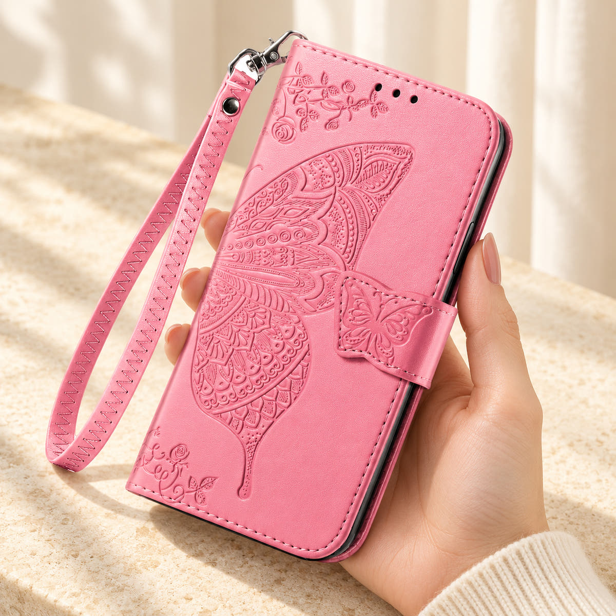Butterfly embossed iPhone wallet case in Pink, handheld closed-front view with butterfly tab and matching wrist loop.