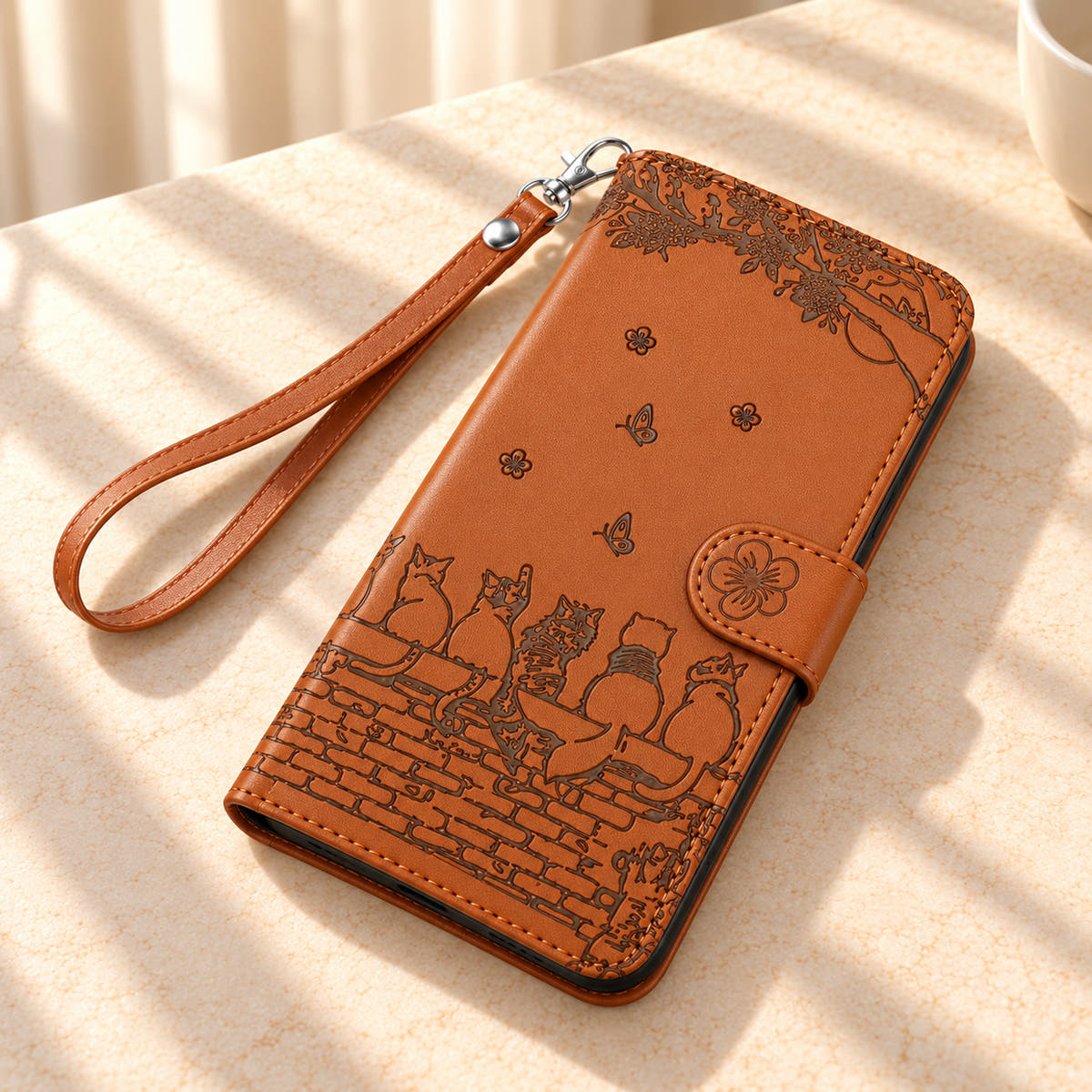 Cognac Brown Cat Garden wallet case and wrist strap on a cream tabletop, showing the flower tab and embossed cats.