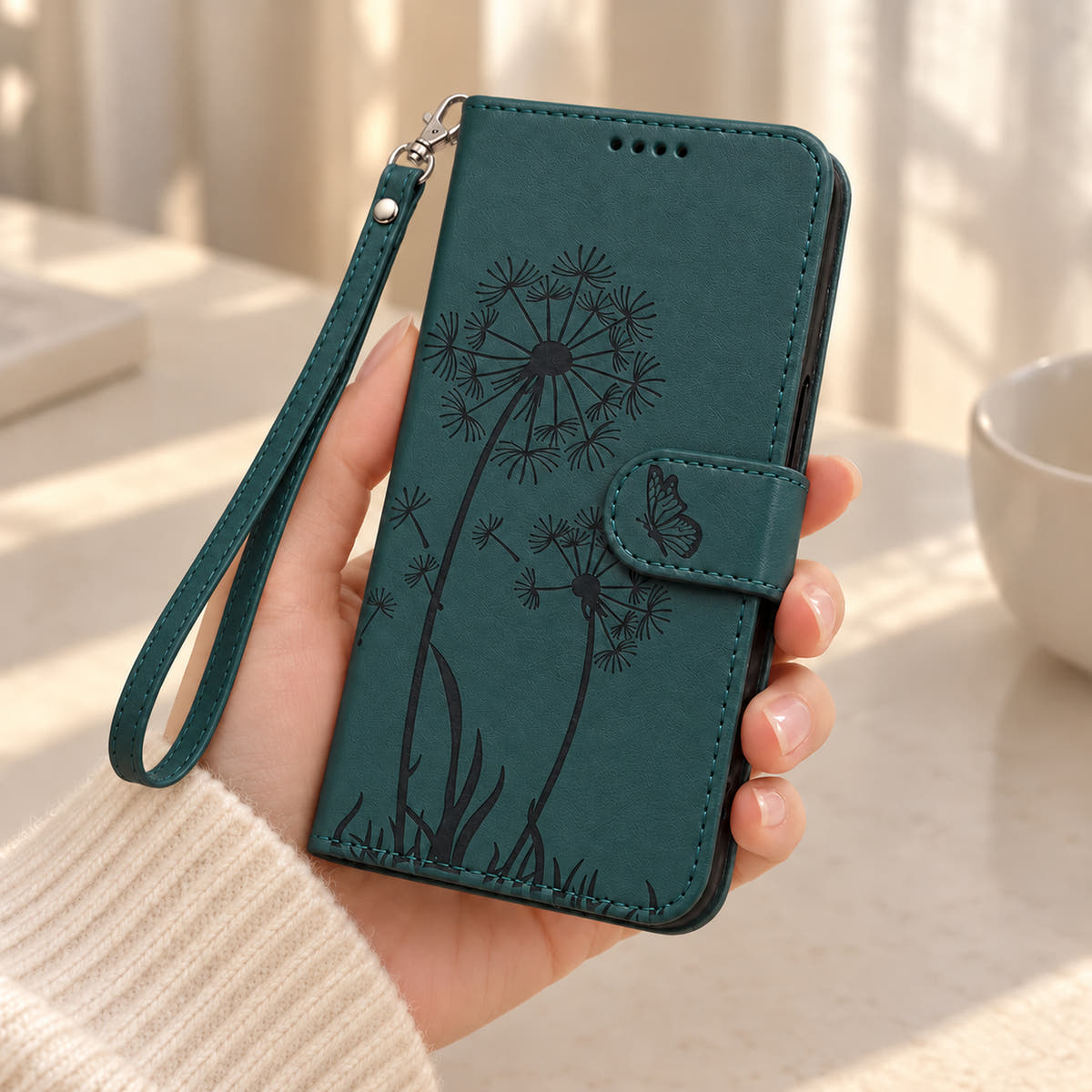 Dark Green dandelion and butterfly wallet phone case held in one hand with a matching wrist strap.