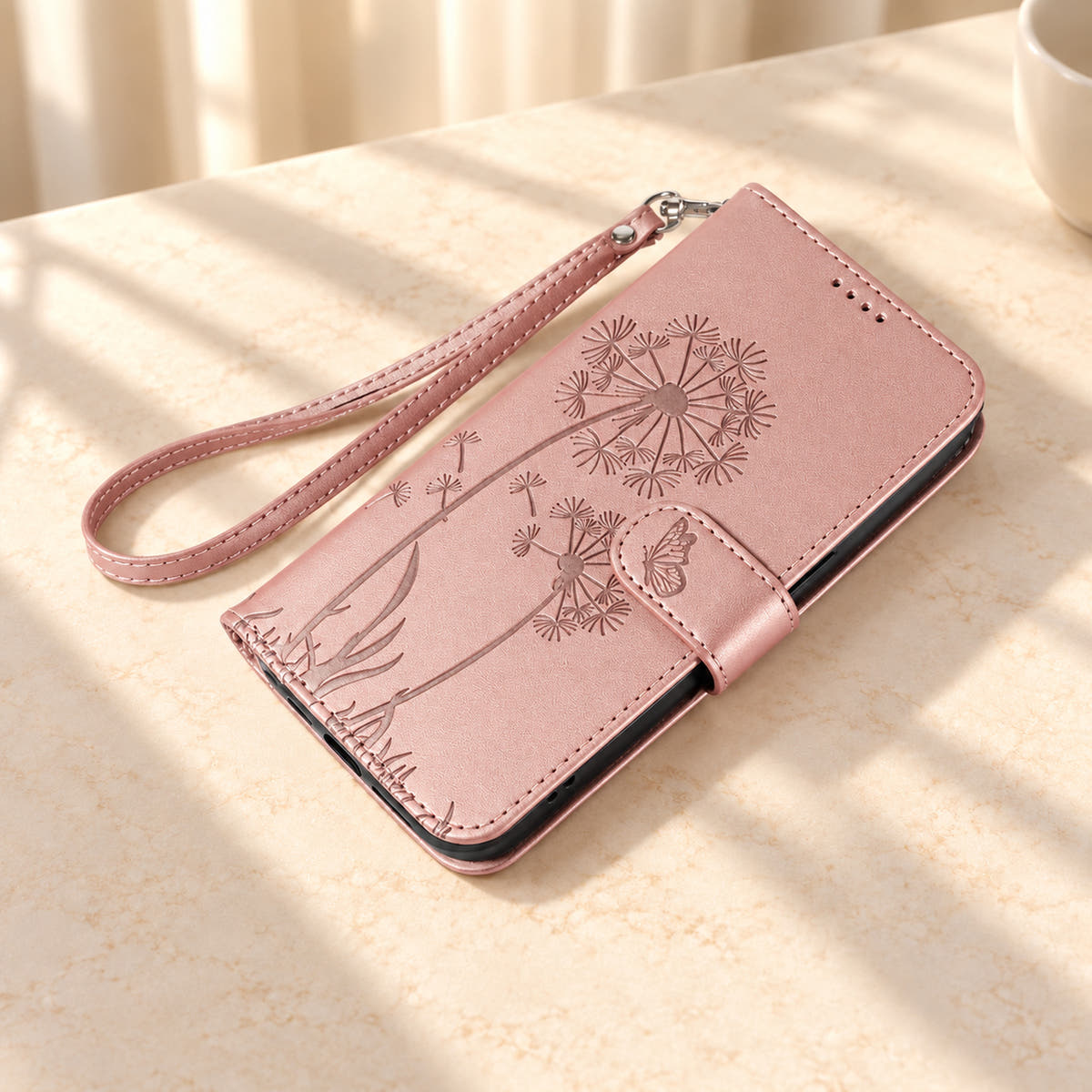 Rose Gold dandelion wallet phone case lying closed with its butterfly tab and matching wrist strap.