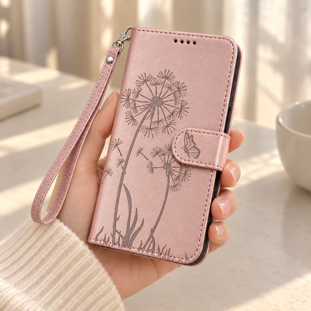 Rose Gold dandelion and butterfly wallet phone case held in one hand with a matching wrist strap.