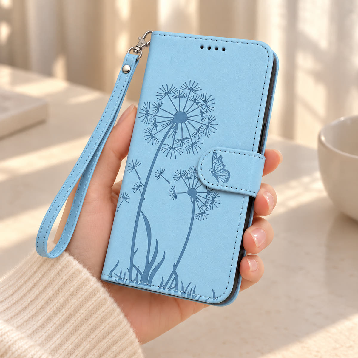 Sky Blue dandelion and butterfly wallet phone case held in one hand with a matching wrist strap.