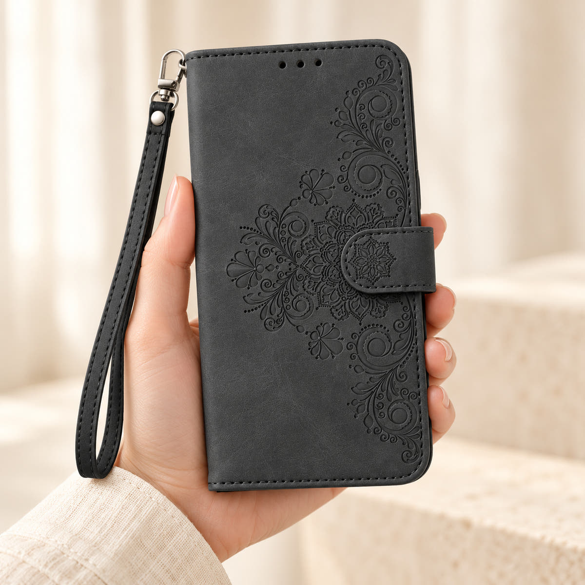 Floral embossed iPhone flip wallet case in Black — closed front held in hand with matching wrist strap