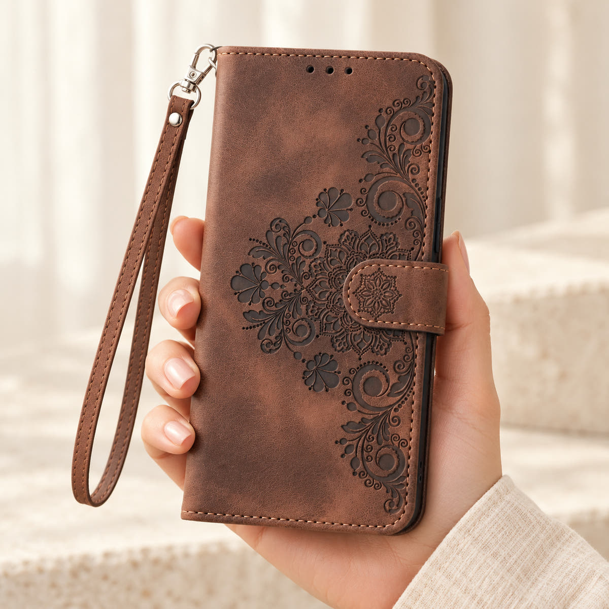 Floral embossed iPhone flip wallet case in Brown — closed front held in hand with matching wrist strap