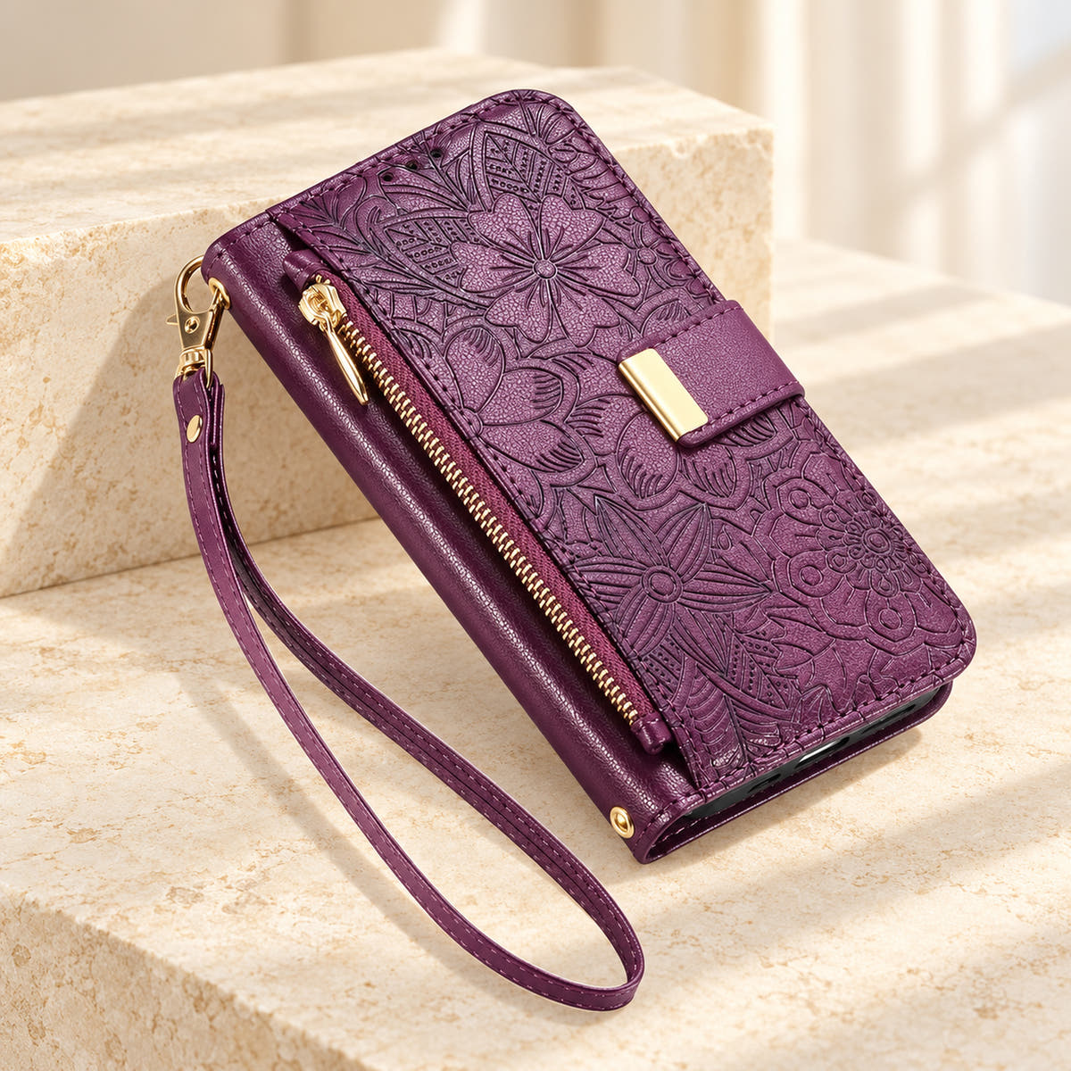 Purple embossed floral iPhone wallet case on a cream surface with the closed zip pouch, gold-tone tab and matching wrist loop
