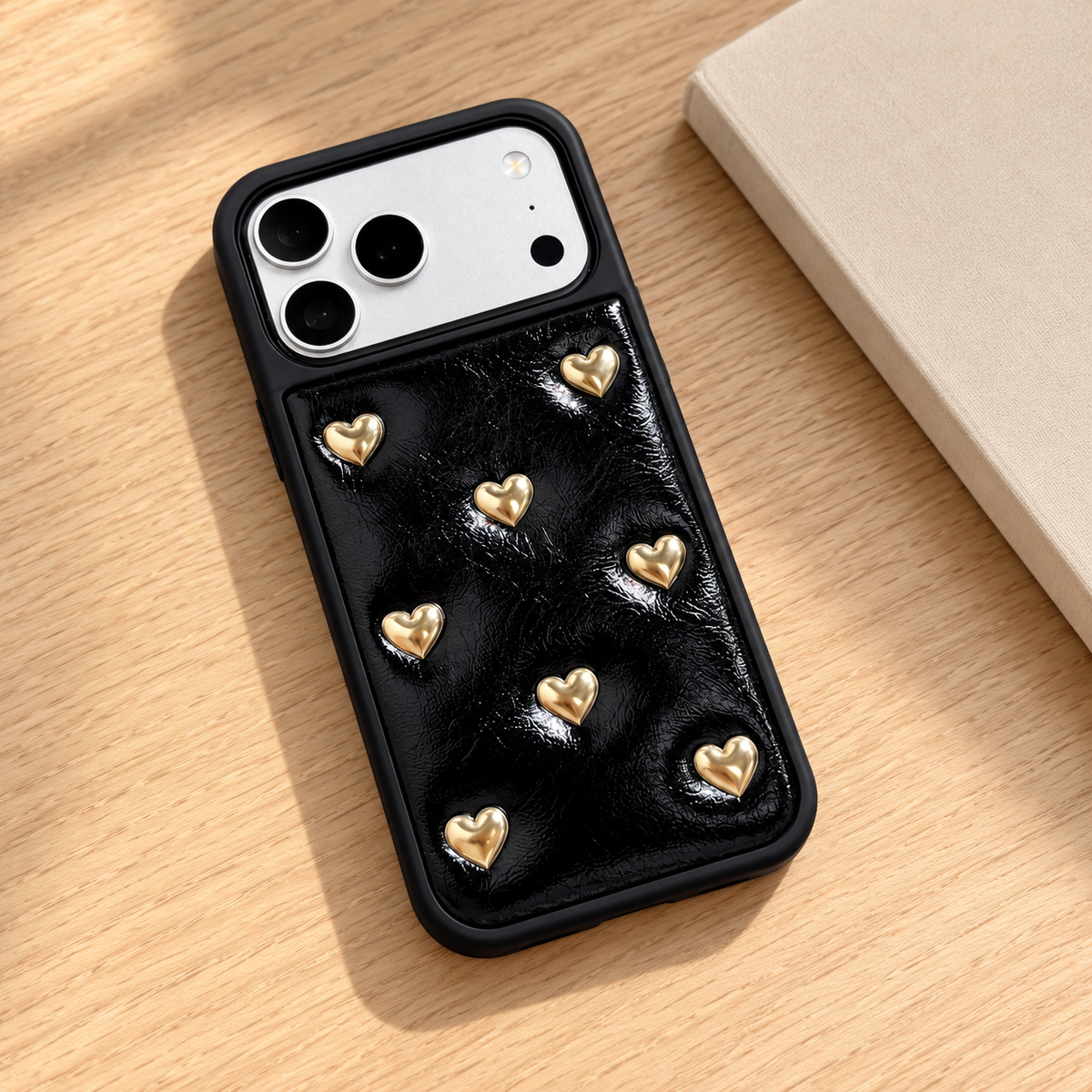 Black iPhone case with raised gold-tone hearts — rear view on an oak desk.