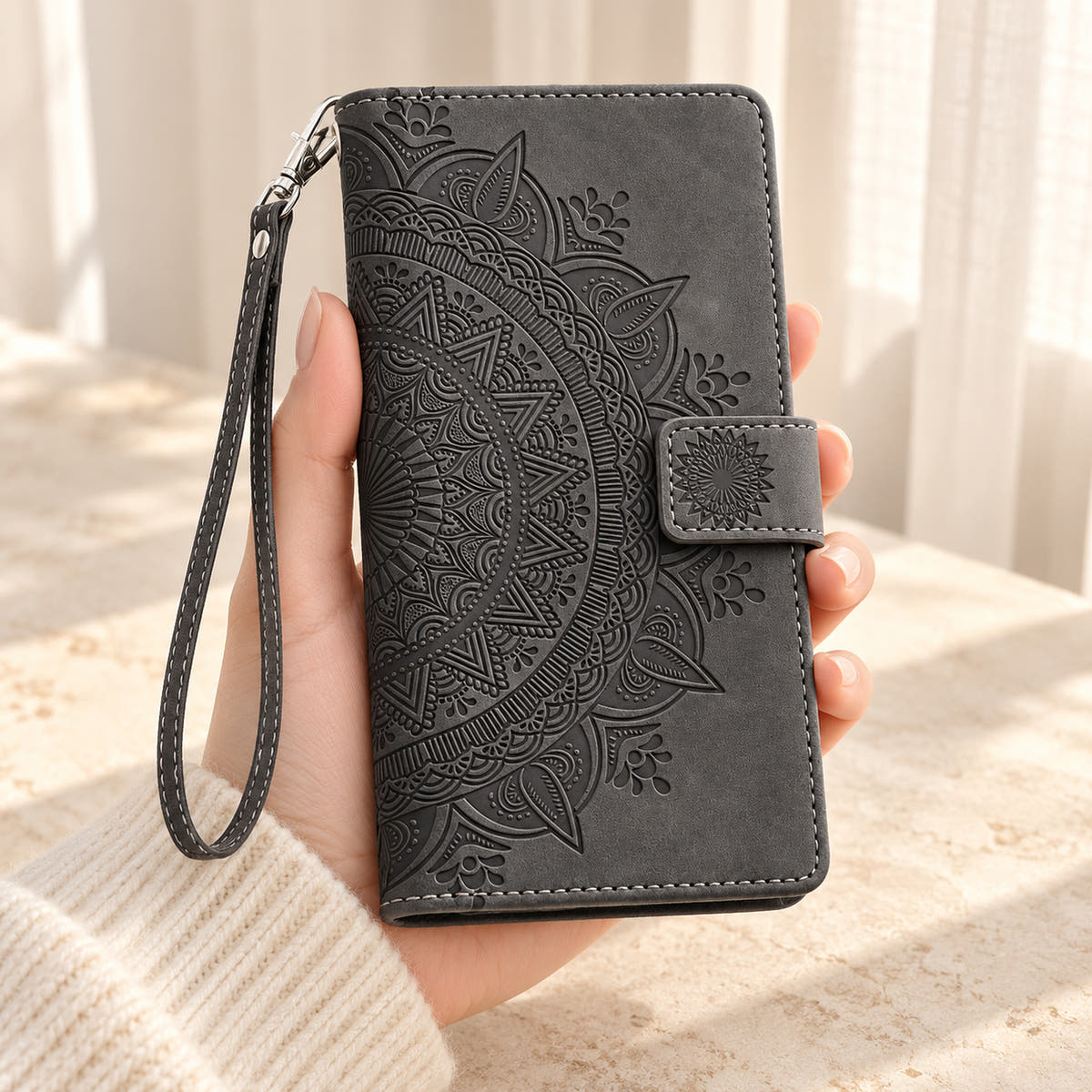 Lotus Mandala iPhone wallet case in Black showing the embossed front and wrist strap held in a cream-sleeve hand