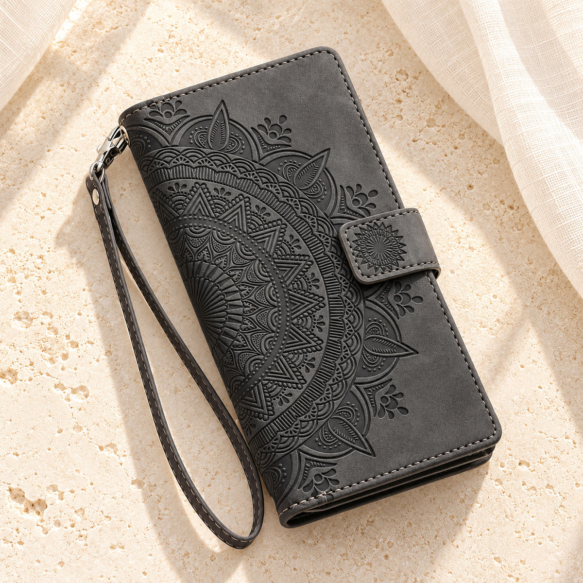 Lotus Mandala iPhone wallet case in Black showing the closed embossed cover and matching wrist loop on limestone