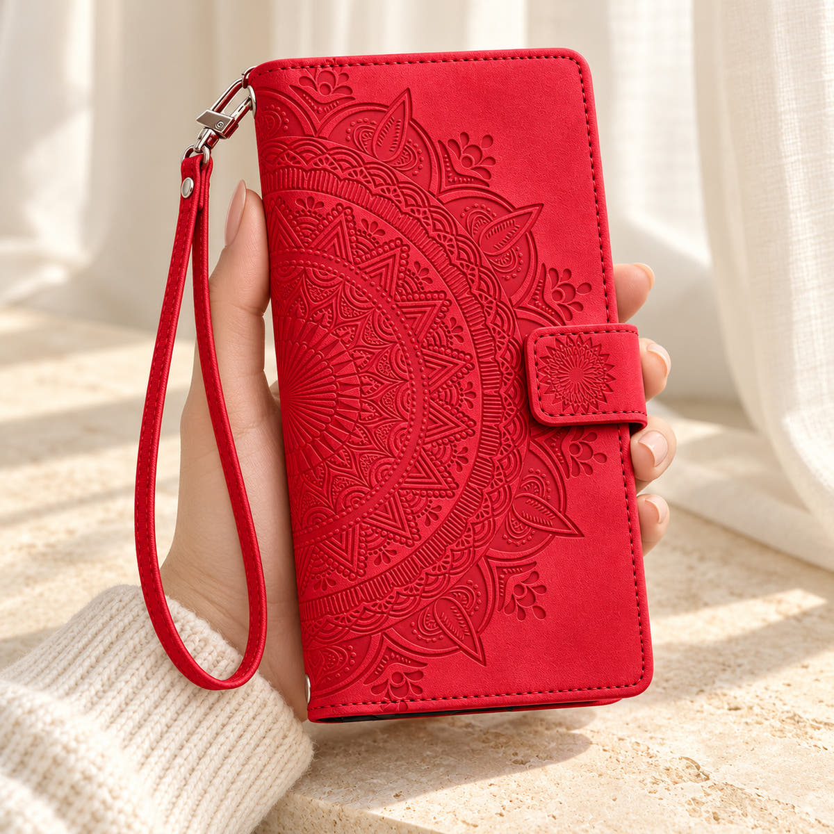 Lotus Mandala iPhone wallet case in Red showing the embossed front and wrist strap held in a cream-sleeve hand