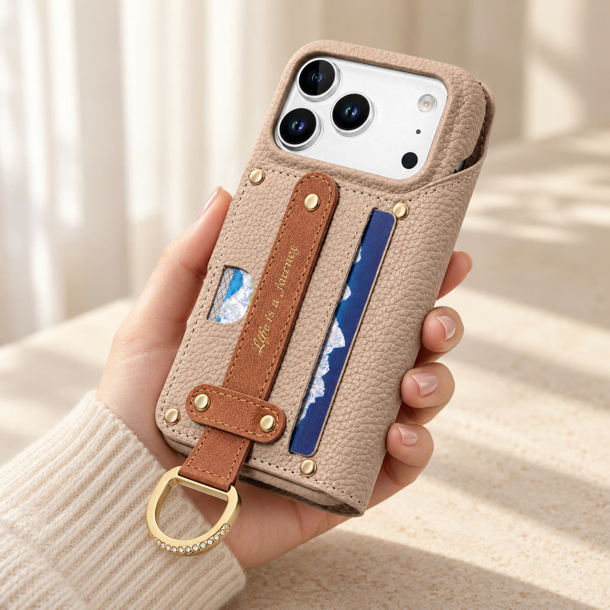 Beige textured iPhone wallet case held in hand, showing the contrasting rear hand strap, card pocket, gold-tone D-ring and camera opening