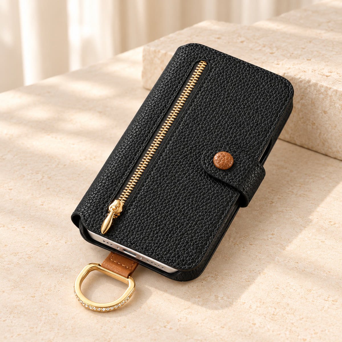 Black textured iPhone wallet case closed on a cream surface, showing the front zip pocket, buttoned tab and attached gold-tone D-ring
