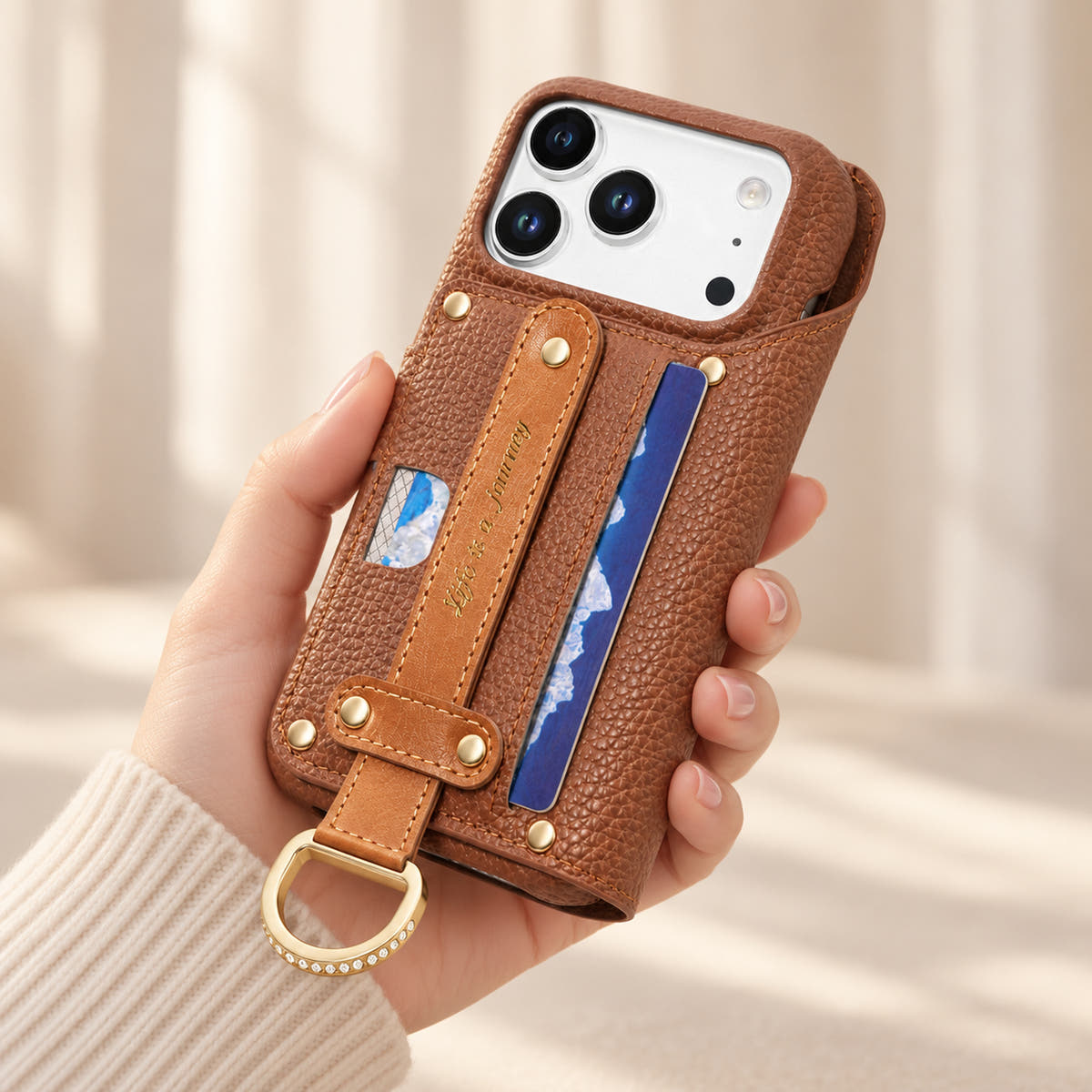 Brown textured iPhone wallet case held in hand, showing the contrasting rear hand strap, card pocket, gold-tone D-ring and camera opening