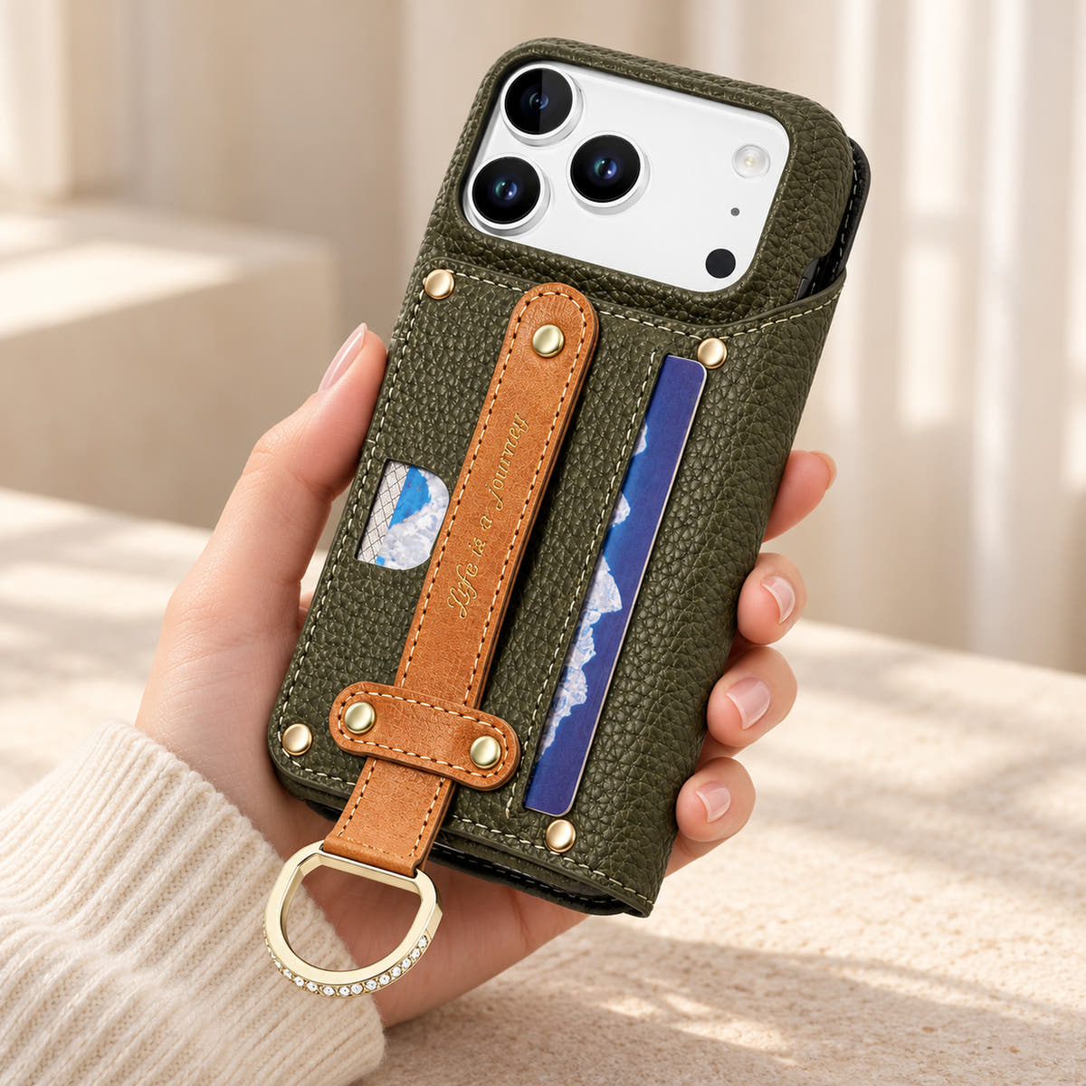 Forest Green textured iPhone wallet case held in hand, showing the contrasting rear hand strap, card pocket, gold-tone D-ring and camera opening