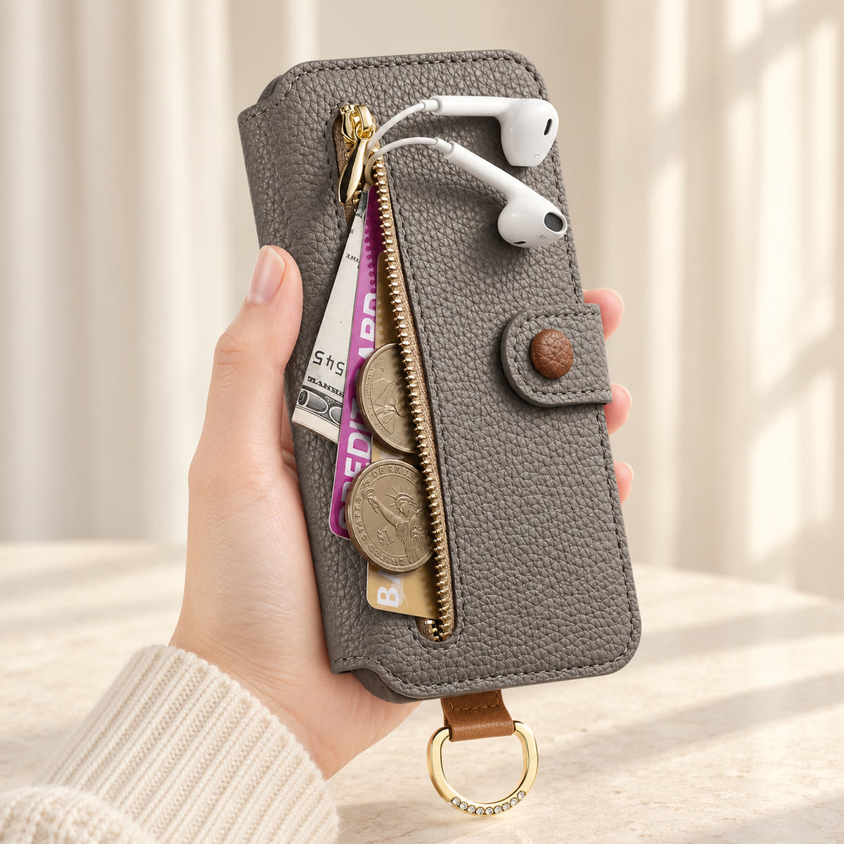 Gray iPhone wallet case with the front zip pocket open, showing illustrated cards, notes, coins and wired earphones beside the buttoned closure