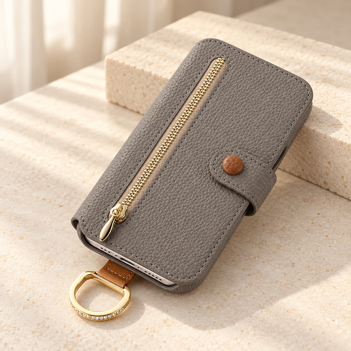 Gray textured iPhone wallet case closed on a cream surface, showing the front zip pocket, buttoned tab and attached gold-tone D-ring