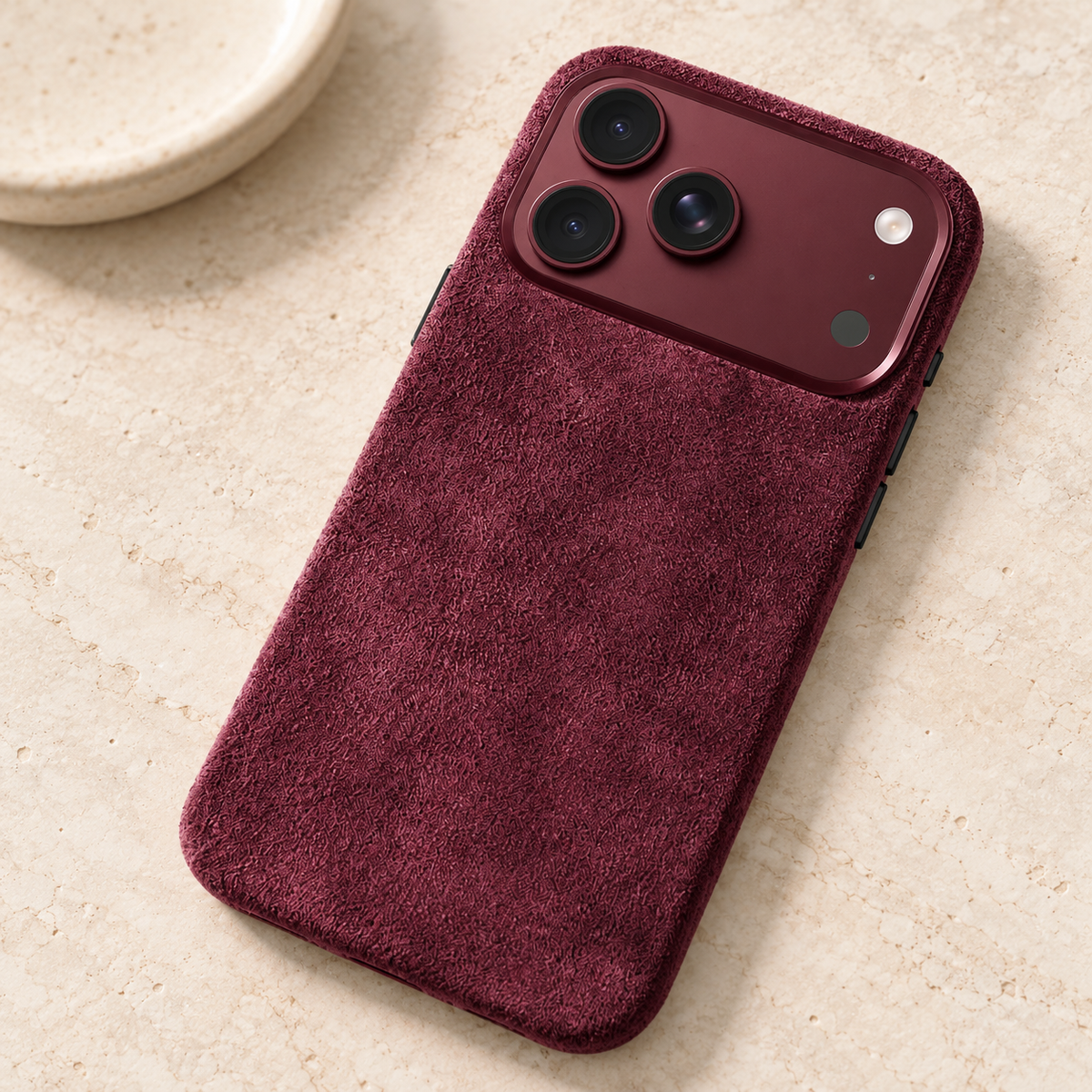Burgundy suede-look textured iPhone case resting on a warm cream stone tabletop.