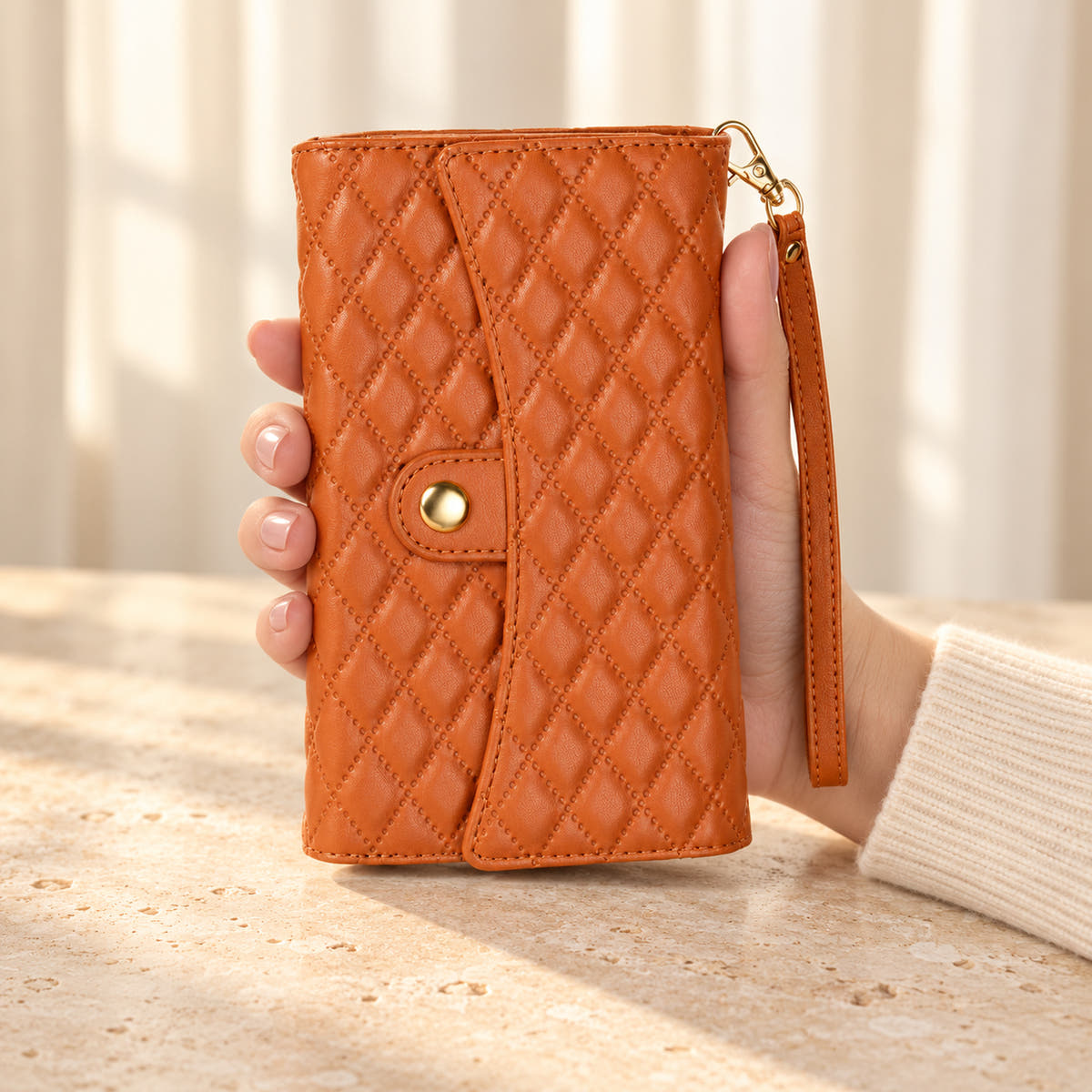 Quilted Zip Wallet iPhone case in Brown held in a cream-sleeved hand, showing its diamond-quilted flap, gold-tone snap and matching wrist strap.