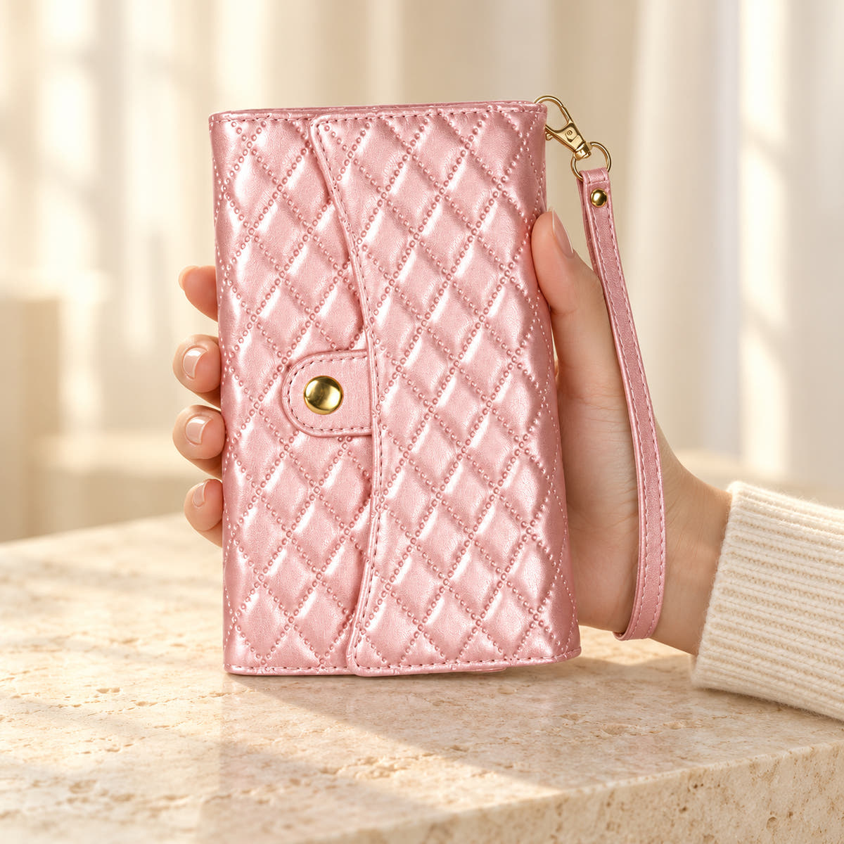 Quilted Zip Wallet iPhone case in Pink held in a cream-sleeved hand, showing its diamond-quilted flap, gold-tone snap and matching wrist strap.