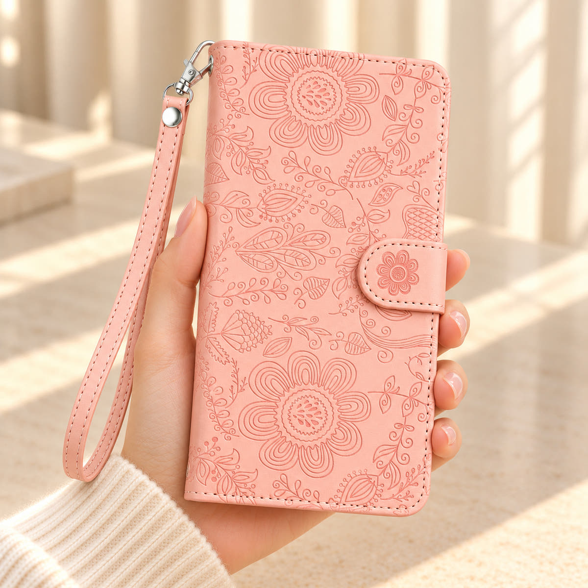 ScarletBloom Pink wallet phone case held in a cream-sleeved hand, showing the floral and paisley embossed cover, flower-decorated tab and matching single-rivet wrist loop.