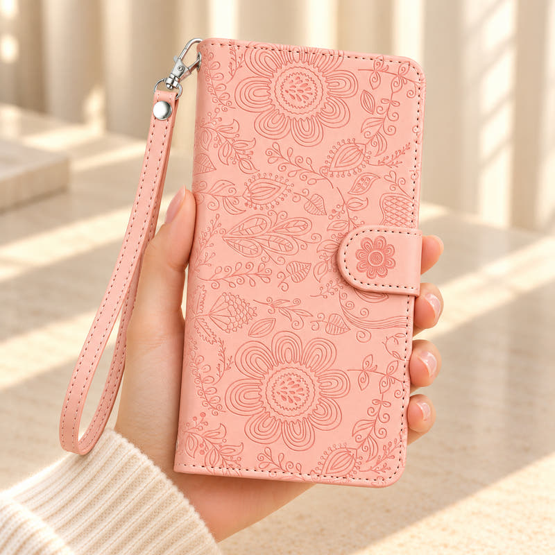 ScarletBloom Pink wallet phone case held in a cream-sleeved hand, showing the floral and paisley embossed cover, flower-decorated tab and matching single-rivet wrist loop.