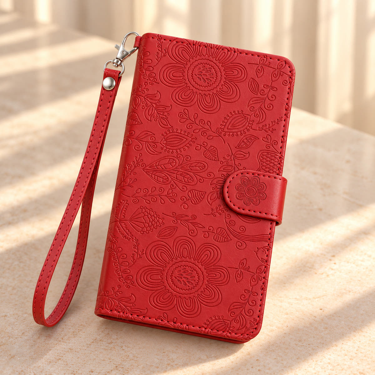 ScarletBloom Red wallet phone case on a sunlit cream limestone tabletop, showing the embossed floral cover and matching wrist loop.