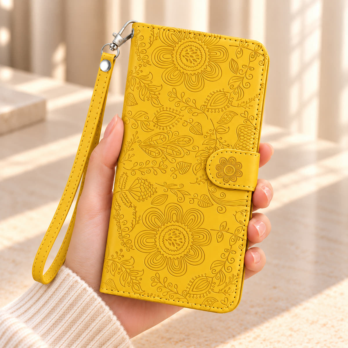 Yellow Scarlet Bloom floral wallet phone case held in one hand, showing its embossed cover, flower-decorated closure tab and matching wrist strap.