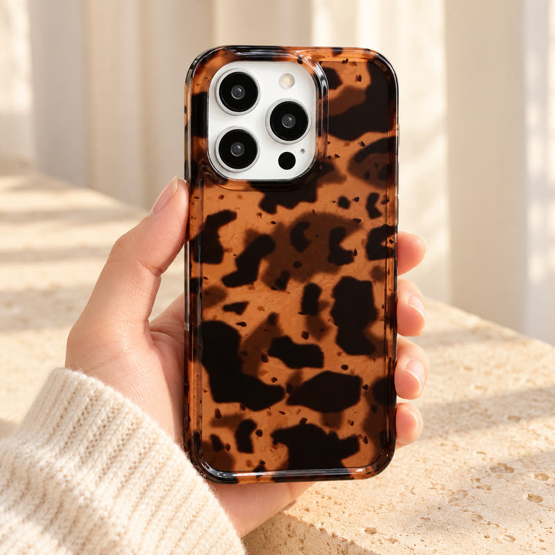 Sienna Luxe Brown tortoiseshell iPhone case held in a cream-sleeved hand, showing its amber-brown pattern, glossy back and clear rim.