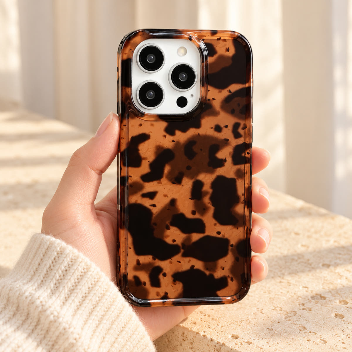 Sienna Luxe Brown tortoiseshell iPhone case held in a cream-sleeved hand, showing its amber-brown pattern, glossy back and clear rim.