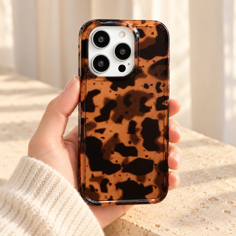 Sienna Luxe Brown tortoiseshell iPhone case held in a cream-sleeved hand, showing its amber-brown pattern, glossy back and clear rim.