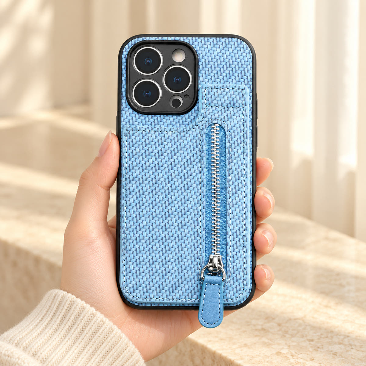 Signature Blue iPhone wallet case held in a cream-sleeved hand, showing its textured rear panel and zip pocket.