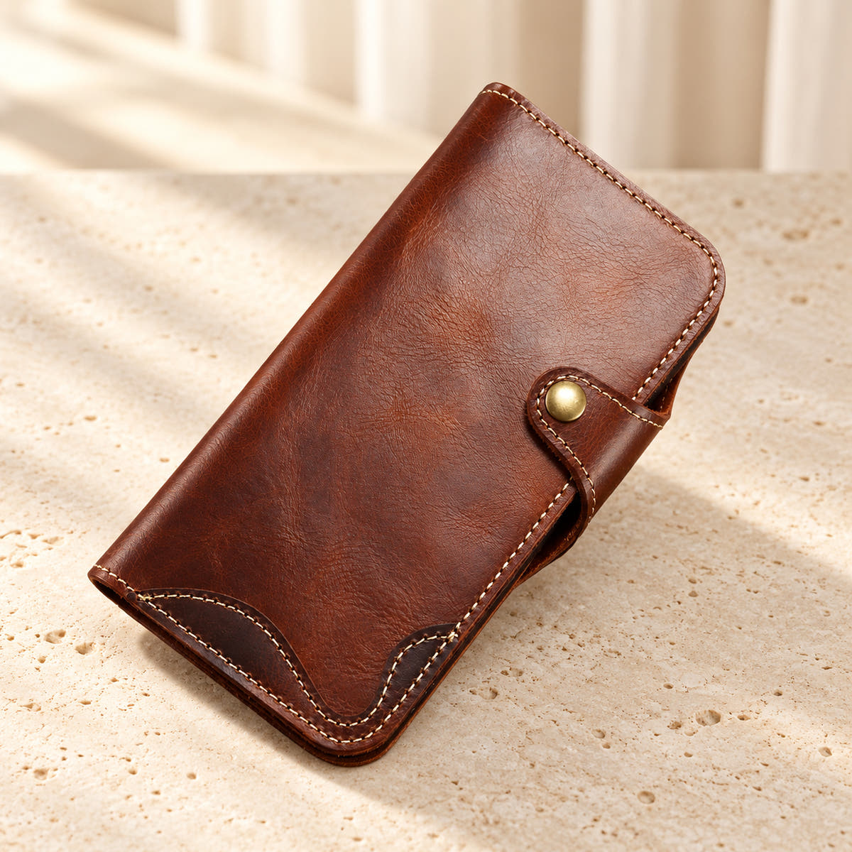 Brown stitched iPhone wallet case resting on cream limestone with its front folio and button closure visible