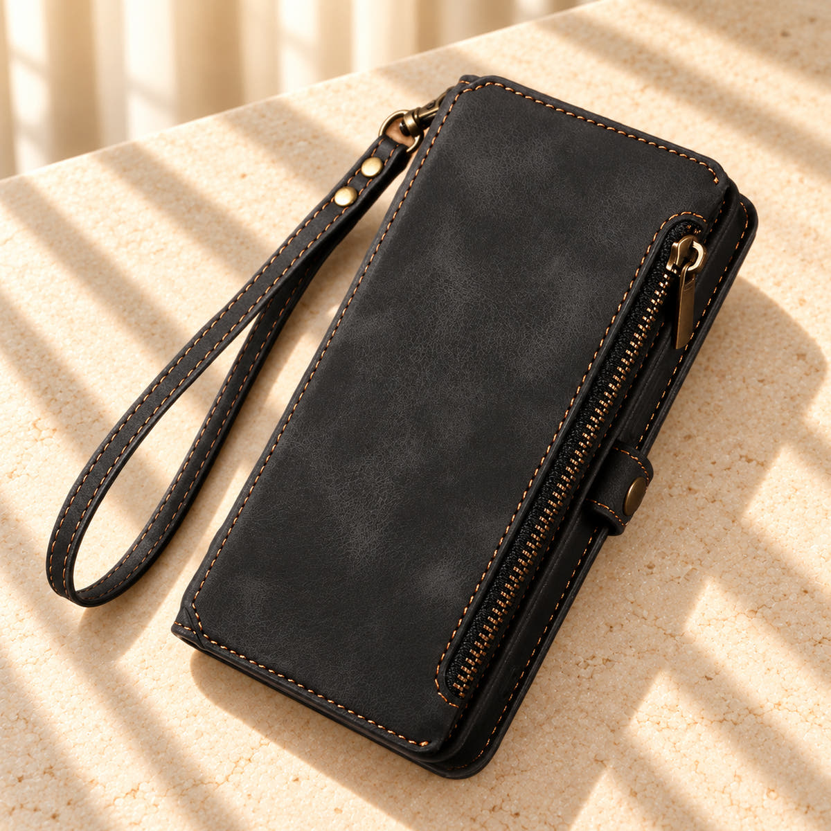BlushCarry Black wallet phone case on a sunlit cream tabletop, showing its straight edge zip pocket and matching wrist strap.