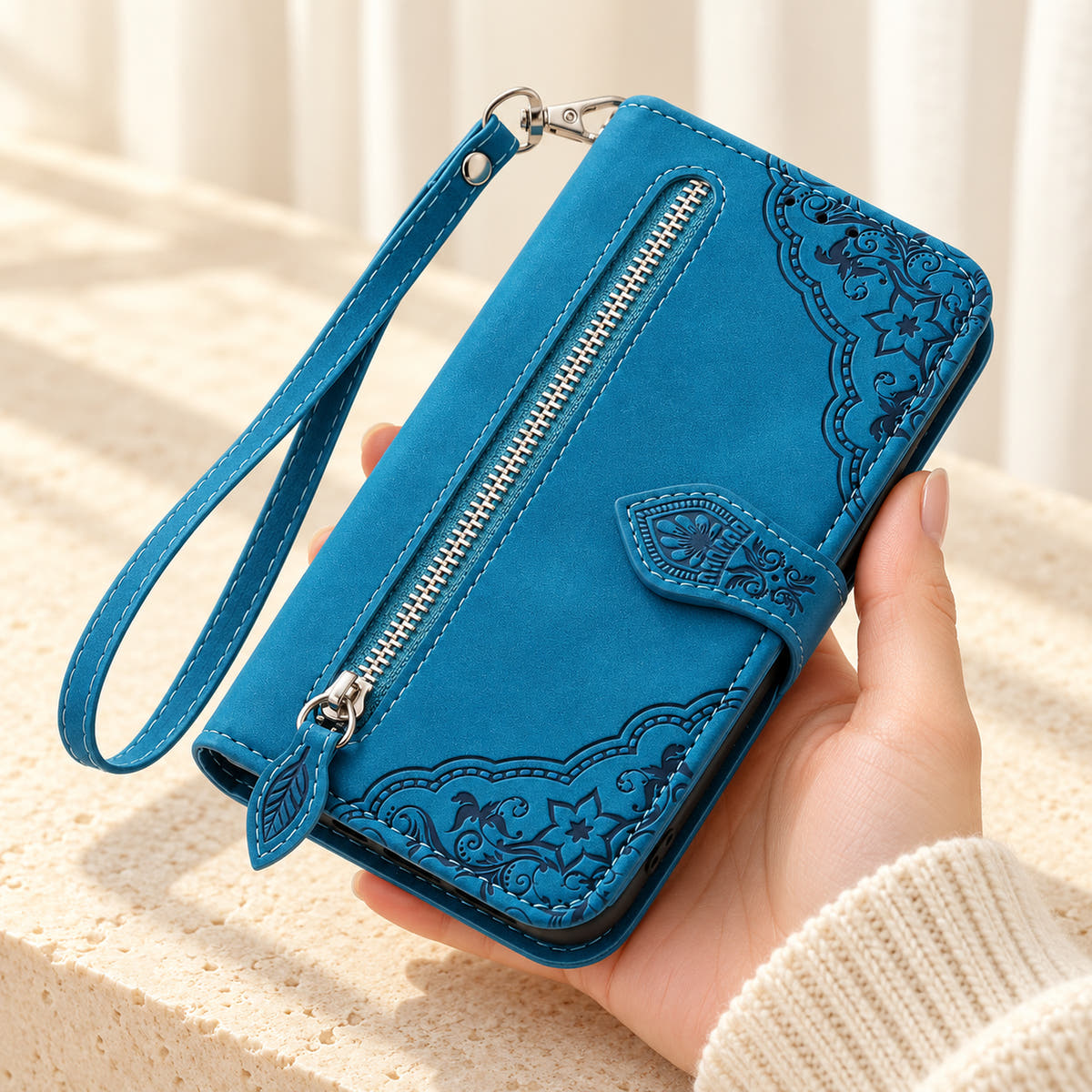 Botanical embossed iPhone wallet case in Blue, handheld front view with silver zip pocket, leaf-shaped pull and matching wrist loop.