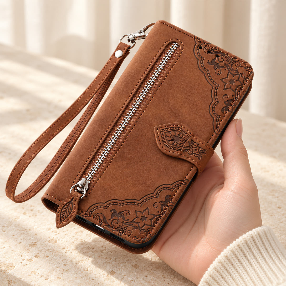 Botanical embossed iPhone wallet case in Brown, handheld front view with silver zip pocket, leaf-shaped pull and matching wrist loop.