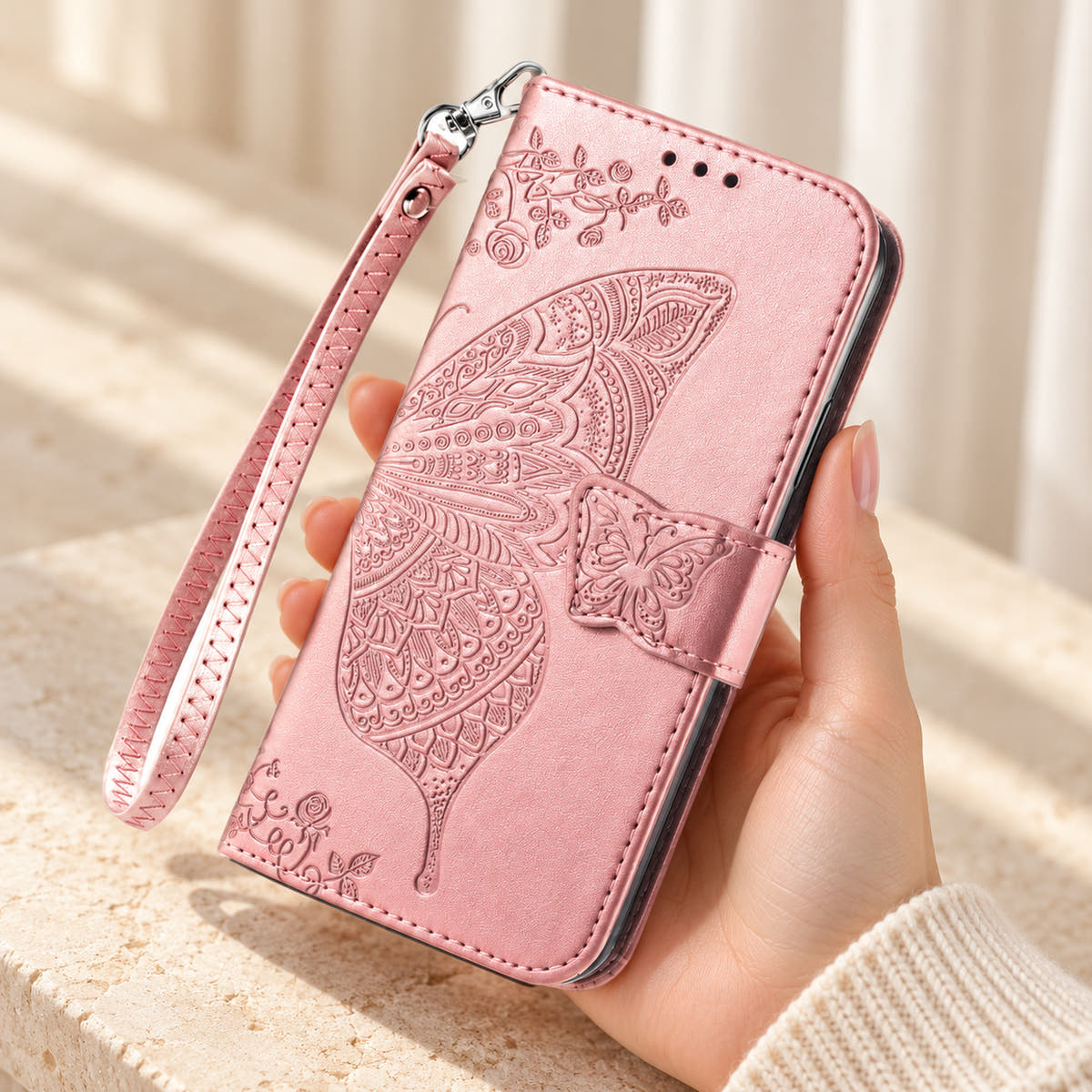 Butterfly embossed iPhone wallet case in Baby Pink, handheld closed-front view with butterfly tab and matching wrist loop.