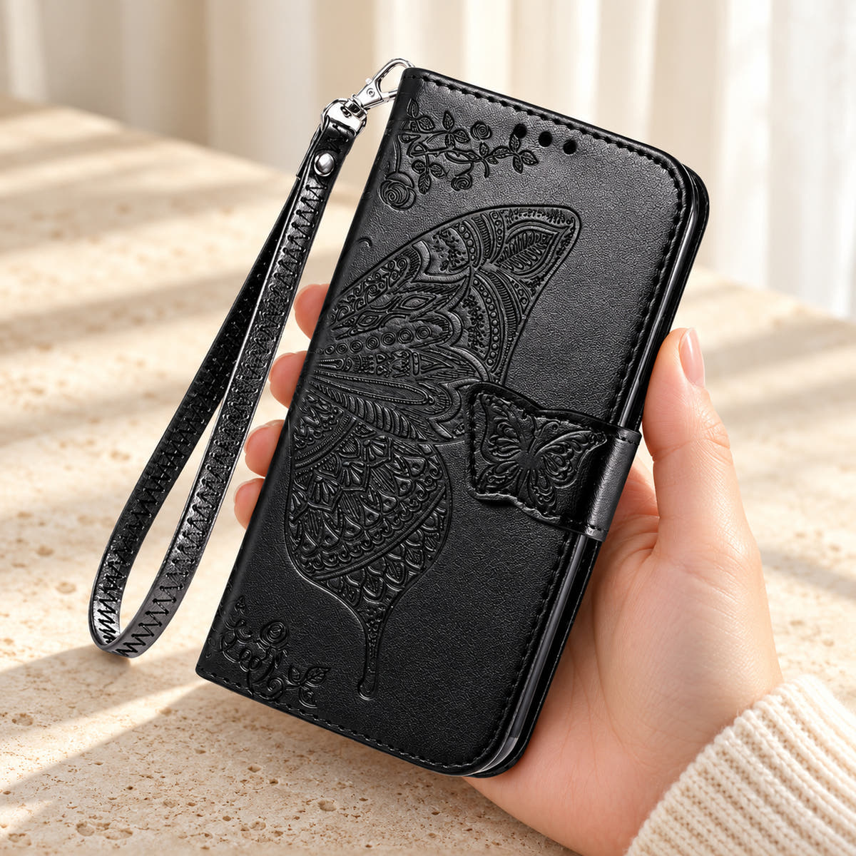 Butterfly embossed iPhone wallet case in Black, handheld closed-front view with butterfly tab and matching wrist loop.