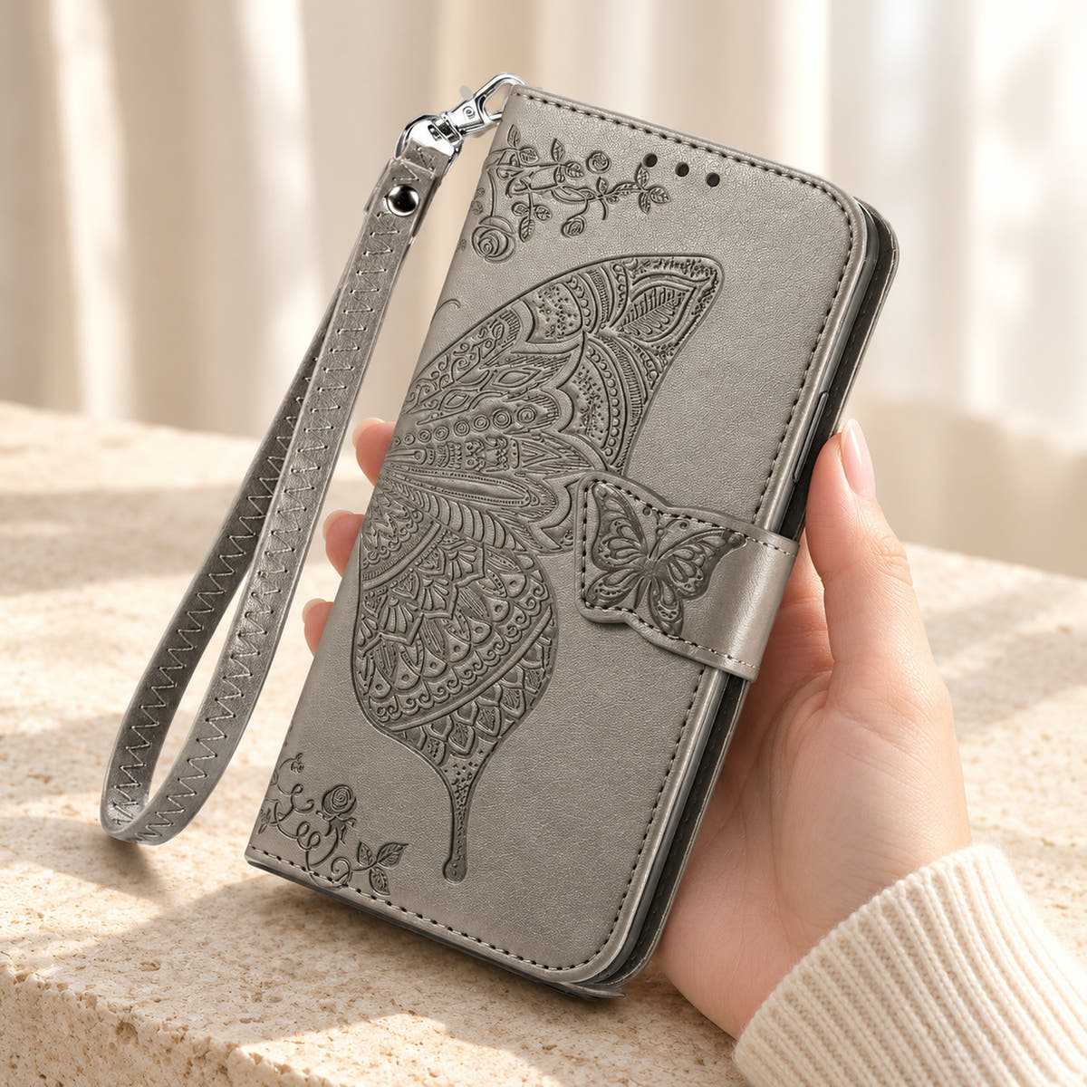 Butterfly embossed iPhone wallet case in Gray, handheld closed-front view with butterfly tab and matching wrist loop.