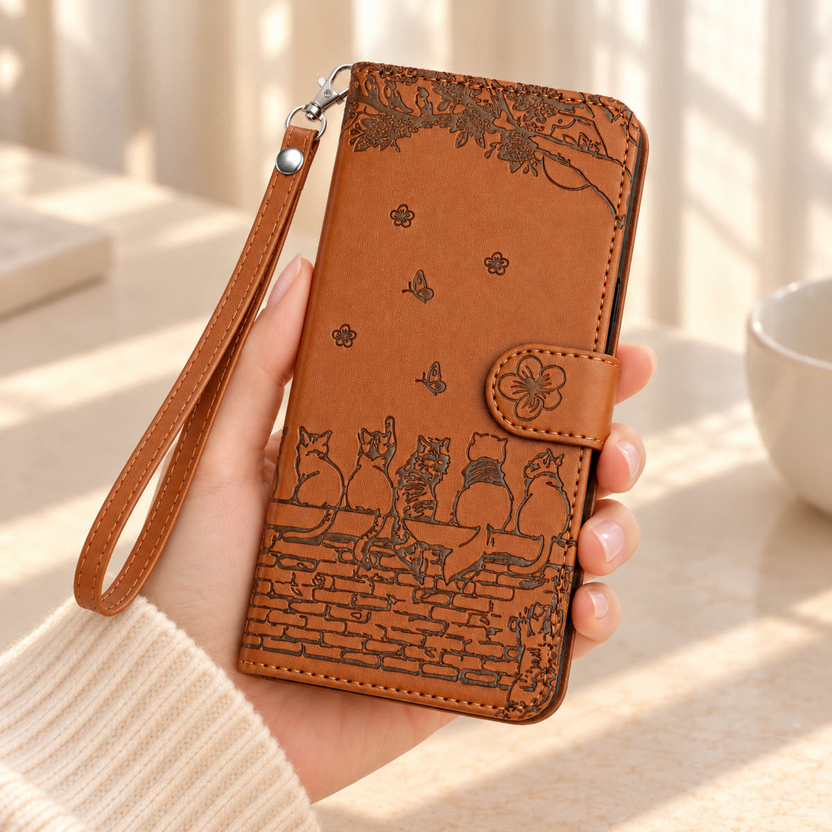 Cognac Brown Cat Garden case held in one hand, showing the embossed cat design, flower closure and wrist strap.