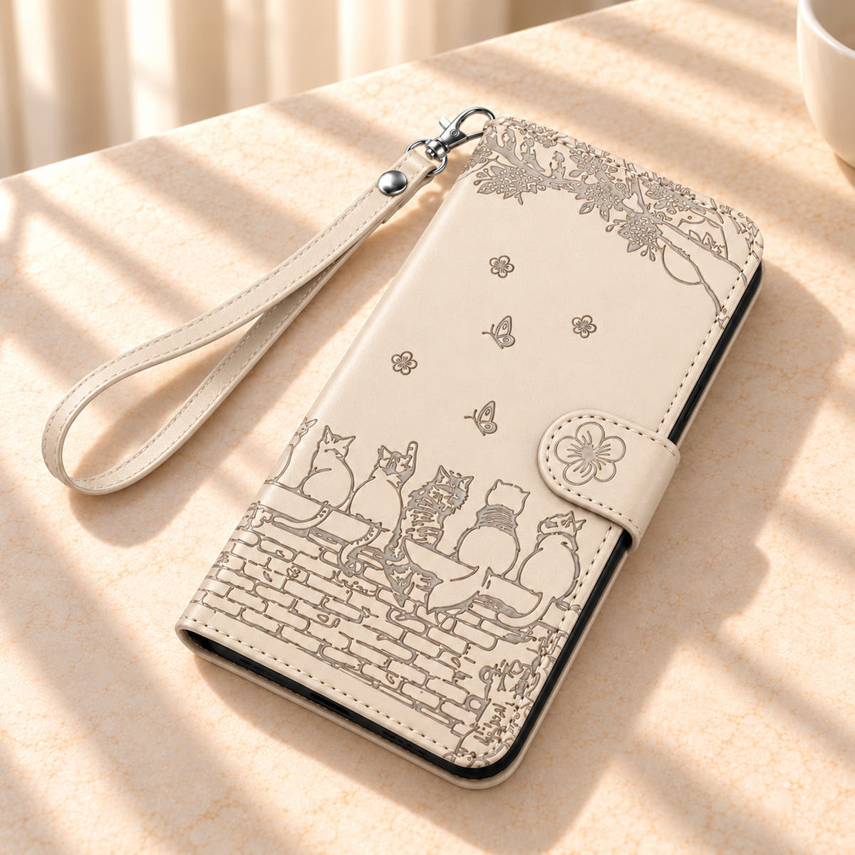 Ivory Cat Garden wallet case and matching wrist strap on a cream tabletop, showing the embossed design.