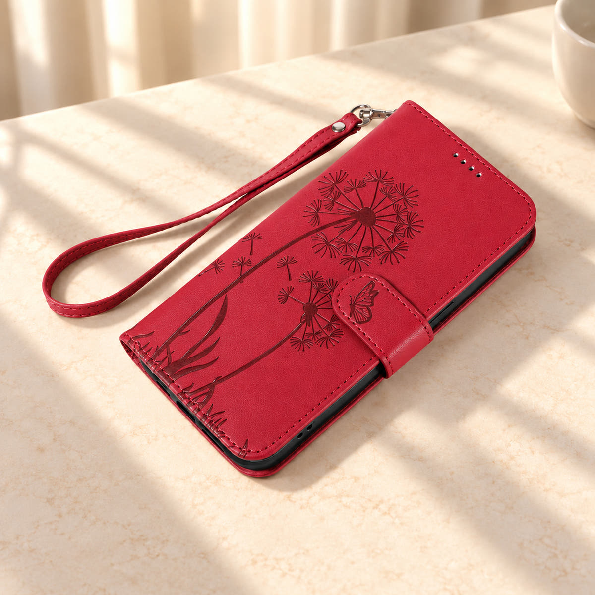 Red dandelion wallet phone case lying closed on cream stone beside its matching wrist strap.