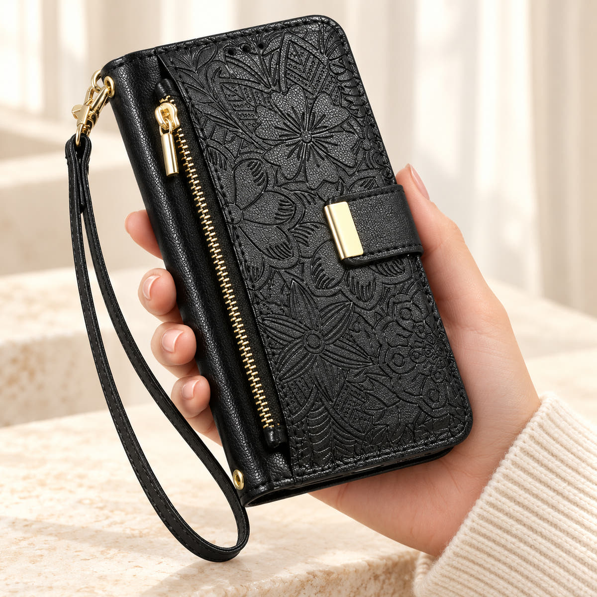 Black floral iPhone wallet case held in hand, showing embossed flowers, gold-tone closure, front zip pouch and matching wrist strap
