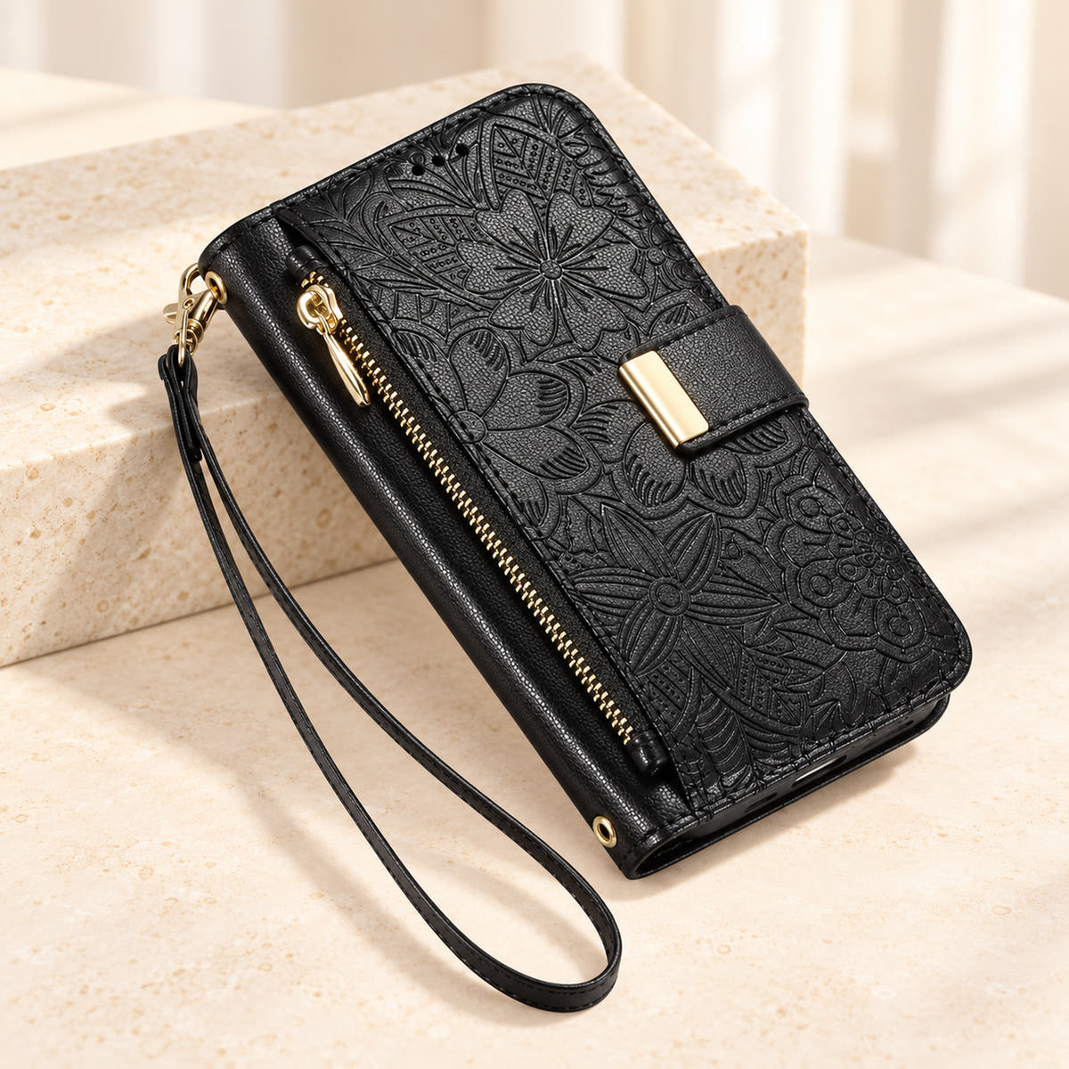 Black embossed floral iPhone wallet case on a cream surface with the closed zip pouch, gold-tone tab and matching wrist loop