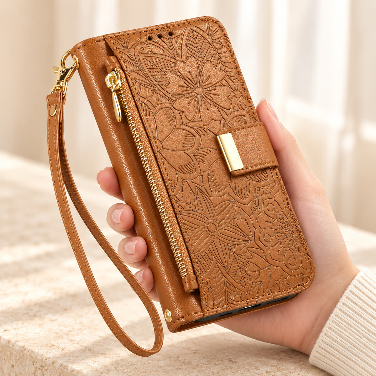 Brown floral iPhone wallet case held in hand, showing embossed flowers, gold-tone closure, front zip pouch and matching wrist strap