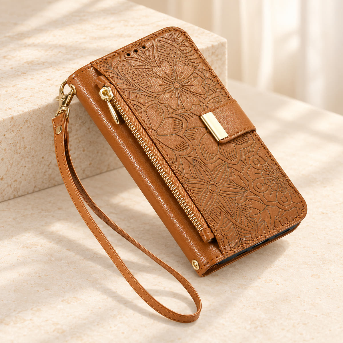 Brown embossed floral iPhone wallet case on a cream surface with the closed zip pouch, gold-tone tab and matching wrist loop
