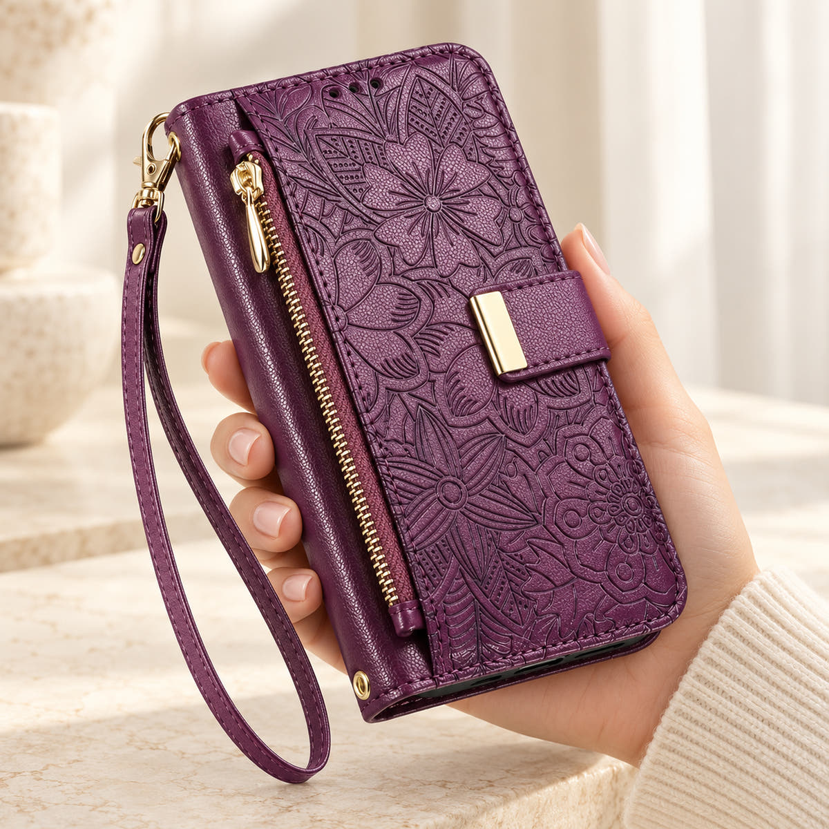 Purple floral iPhone wallet case held in hand, showing embossed flowers, gold-tone closure, front zip pouch and matching wrist strap