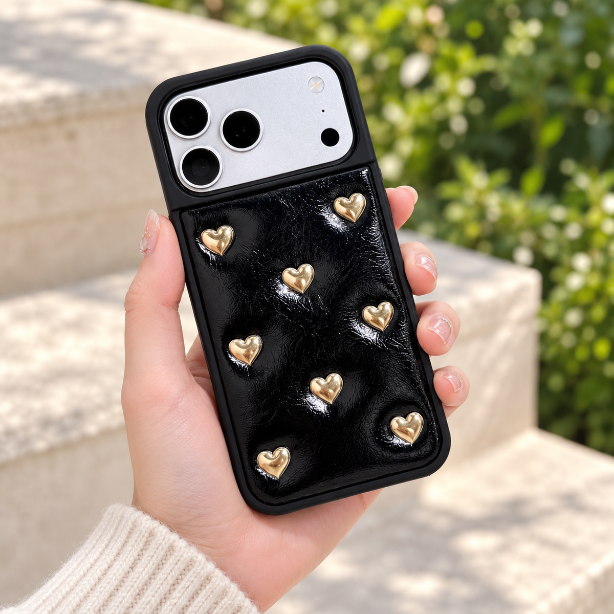 Black iPhone case with raised gold-tone hearts — rear view in daylight.