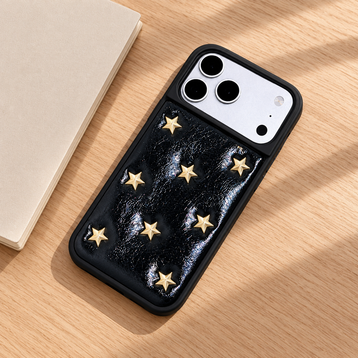 Black iPhone case with raised gold-tone stars — rear view on an oak desk.