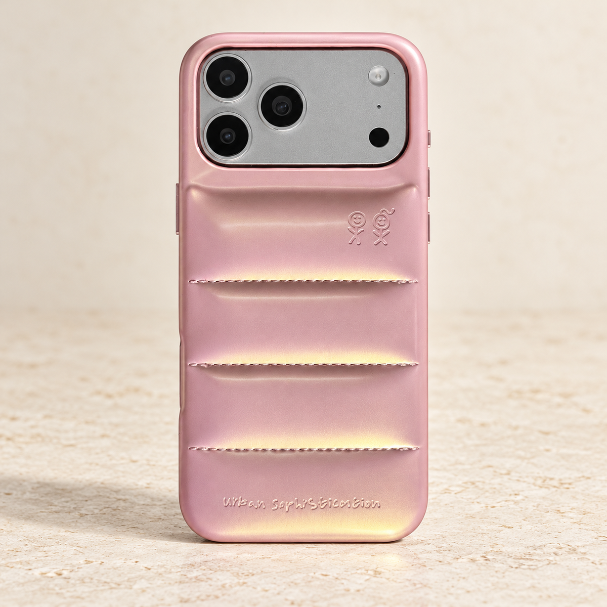 Iridescent Pink puffer iPhone case with three stitched seams, four raised sections and wide camera opening, on warm cream stone.